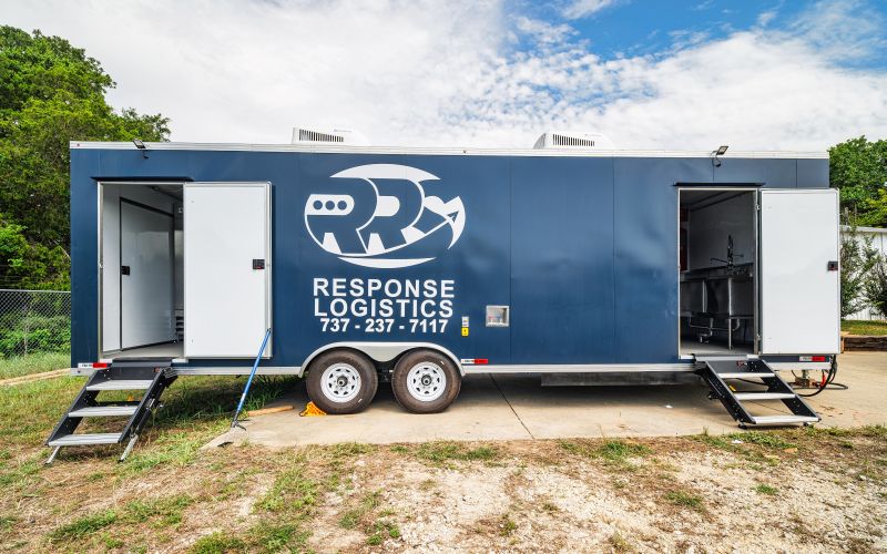 A large white mobile kitchen trailer is parked outdoors on a temporary platform, equipped with ventilation units and propane tanks.